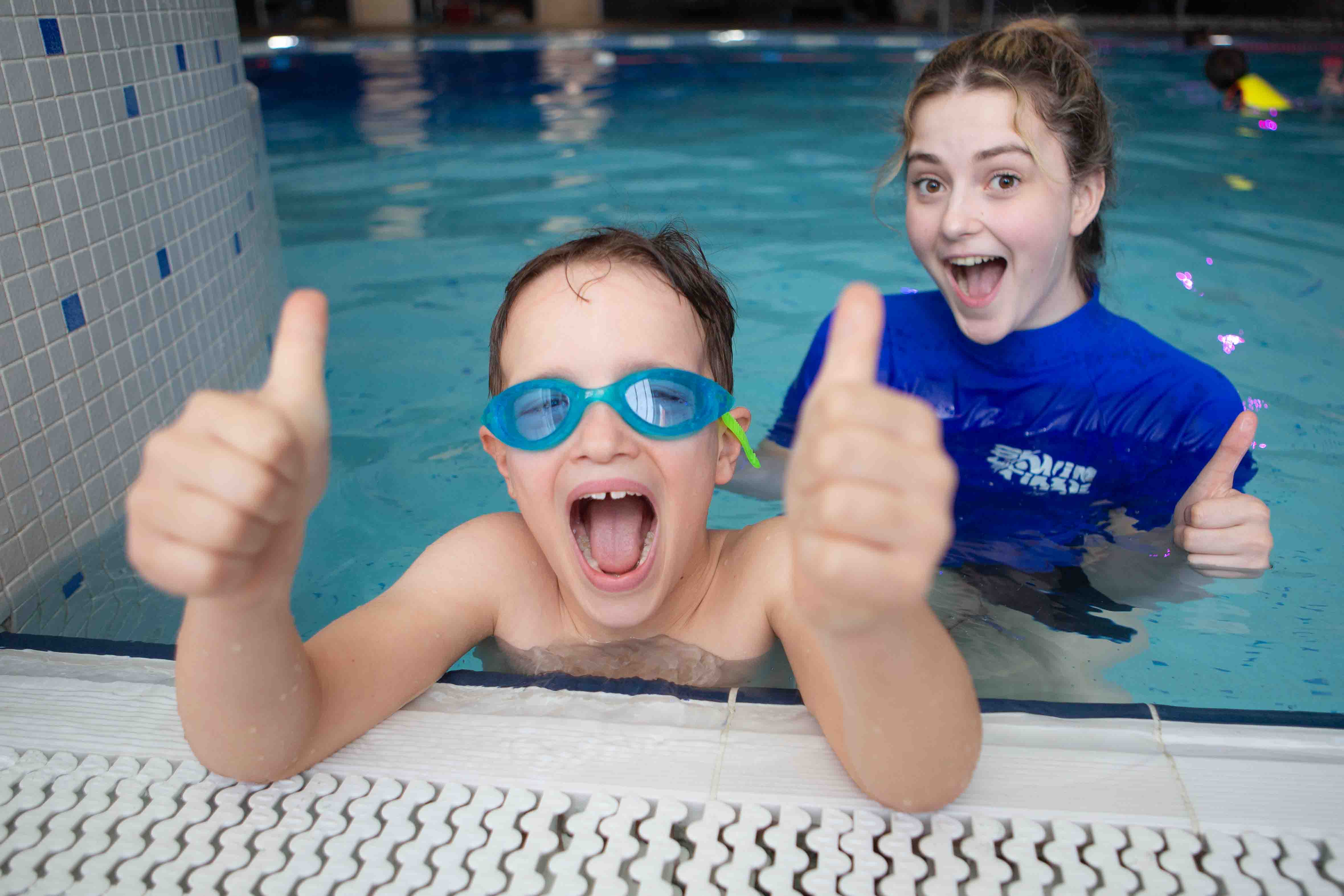 Image of young boy in swimming pool with thumbs up, behind him in the water is a female swimming teacher looking delighted at his progress